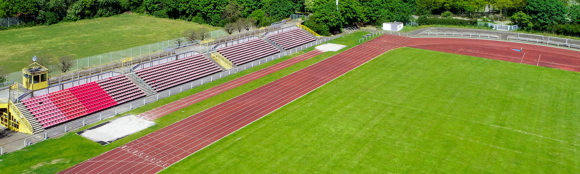 Luftbild des Leichtathletikstadions im Bürgerpark Nord: die rote Laufbahn mit nummerierten Startplätzen, das grüne Innenfeld und die Tribüne mit ihren roten Sitzreihen.